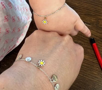 Close-up of adult and child wrists wearing matching silver chain bracelets with small white-and-yellow daisy charms, resting on a dark wooden table with a red pen at the edge.
