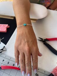 Hand wearing a delicate silver chain bracelet with a small turquoise cross bead on a crafting table with pliers and a cutting mat — DIY jewelry making