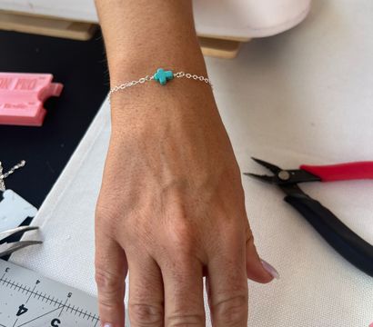 Hand wearing a delicate silver chain bracelet with a small turquoise cross bead on a crafting table with pliers and a cutting mat — DIY jewelry making