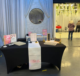 Indoor vendor booth at a craft fair or wedding expo featuring a black table with a white runner displaying minimalist jewelry on gold stands and trays, soft gray draped backdrop with round mirror, and event space with hanging greenery and attendees in the background.