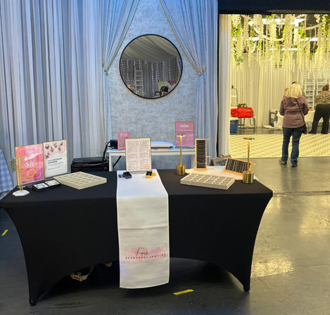 Indoor vendor booth at a craft fair or wedding expo featuring a black table with a white runner displaying minimalist jewelry on gold stands and trays, soft gray draped backdrop with round mirror, and event space with hanging greenery and attendees in the background.