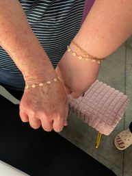 Close-up of two wrists wearing matching gold daisy chain bracelets — a freckled hand and a smoother hand side-by-side against a striped top and a pink ribbed stool in the background.