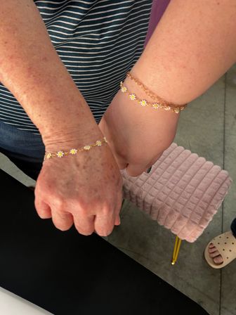 Close-up of two wrists wearing matching gold daisy chain bracelets — a freckled hand and a smoother hand side-by-side against a striped top and a pink ribbed stool in the background.