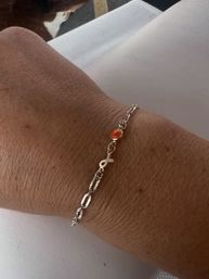 Close-up of a tanned wrist wearing a dainty silver chain bracelet with a small fish charm and an oval orange-red gemstone, resting on white fabric