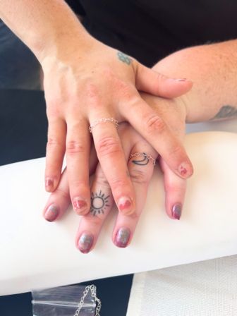 Stacked hands on a white armrest showing chipped metallic rose manicure, sun and crescent finger tattoos, and delicate silver heart-chain rings — playful close-up.