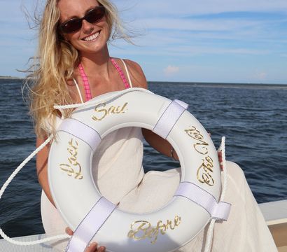Smiling sunlit woman in sunglasses holding a white life preserver aboard a boat on calm coastal waters under a blue summer sky