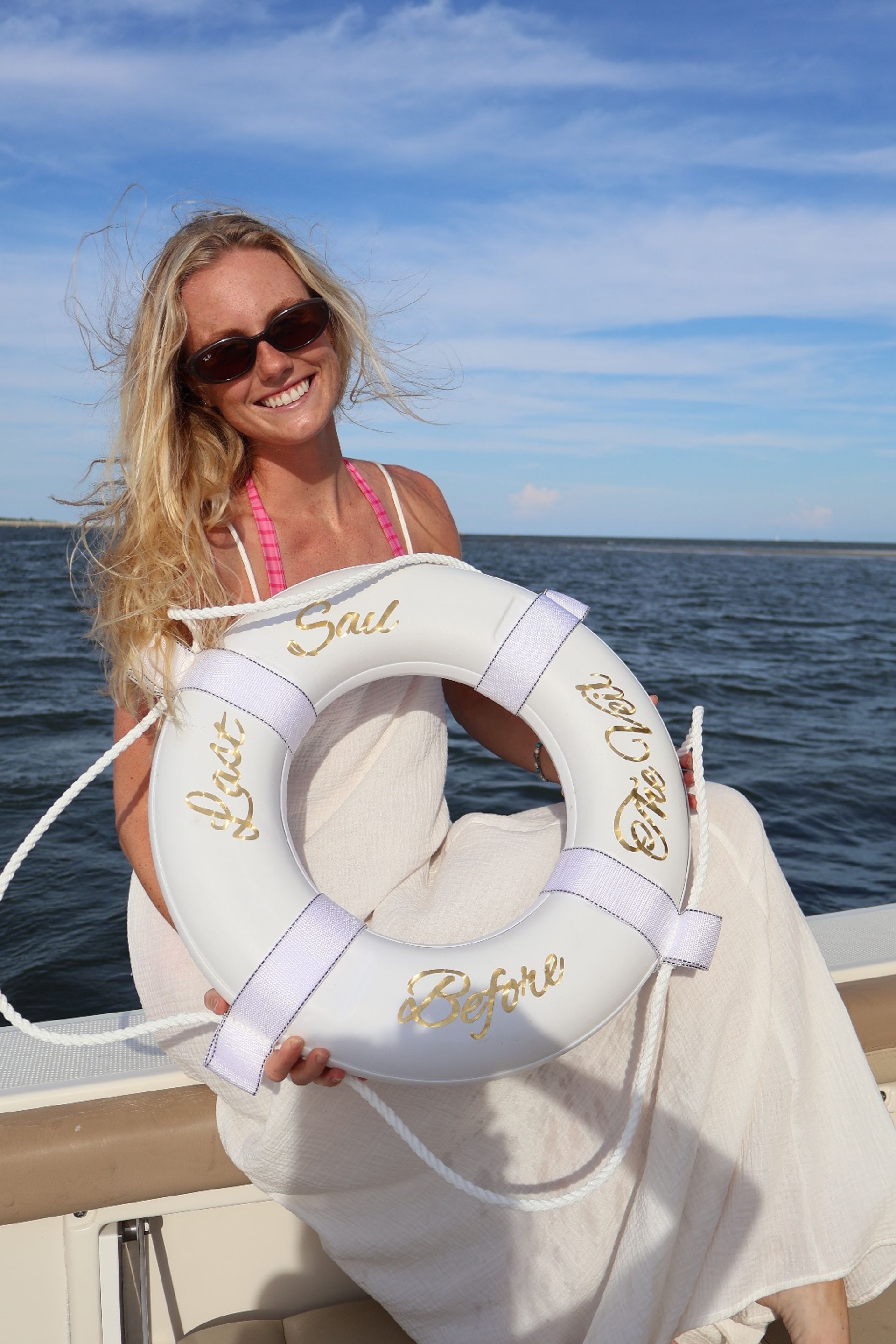 Smiling sunlit woman in sunglasses holding a white life preserver aboard a boat on calm coastal waters under a blue summer sky