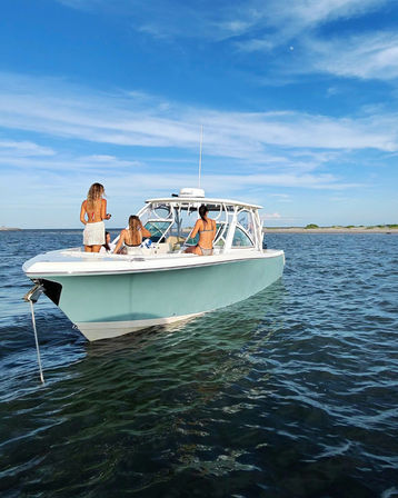 Sun-soaked summer scene: three women in swimsuits relaxing on a light-blue center-console boat anchored off a sandy coastline, calm ocean and clear blue sky.