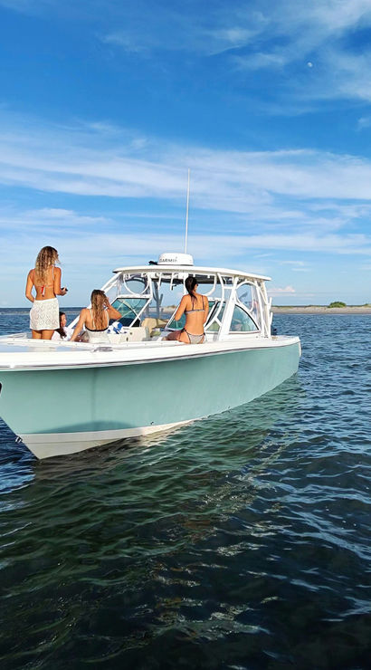 Sun-soaked summer scene: three women in swimsuits relaxing on a light-blue center-console boat anchored off a sandy coastline, calm ocean and clear blue sky.