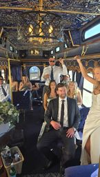 Luxury navy-and-gold party bus interior with ornate chandelier and velvet seating, a cheerful wedding party in formal attire including a bride in a white gown and guests enjoying drinks.