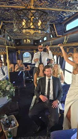Luxury navy-and-gold party bus interior with ornate chandelier and velvet seating, a cheerful wedding party in formal attire including a bride in a white gown and guests enjoying drinks.
