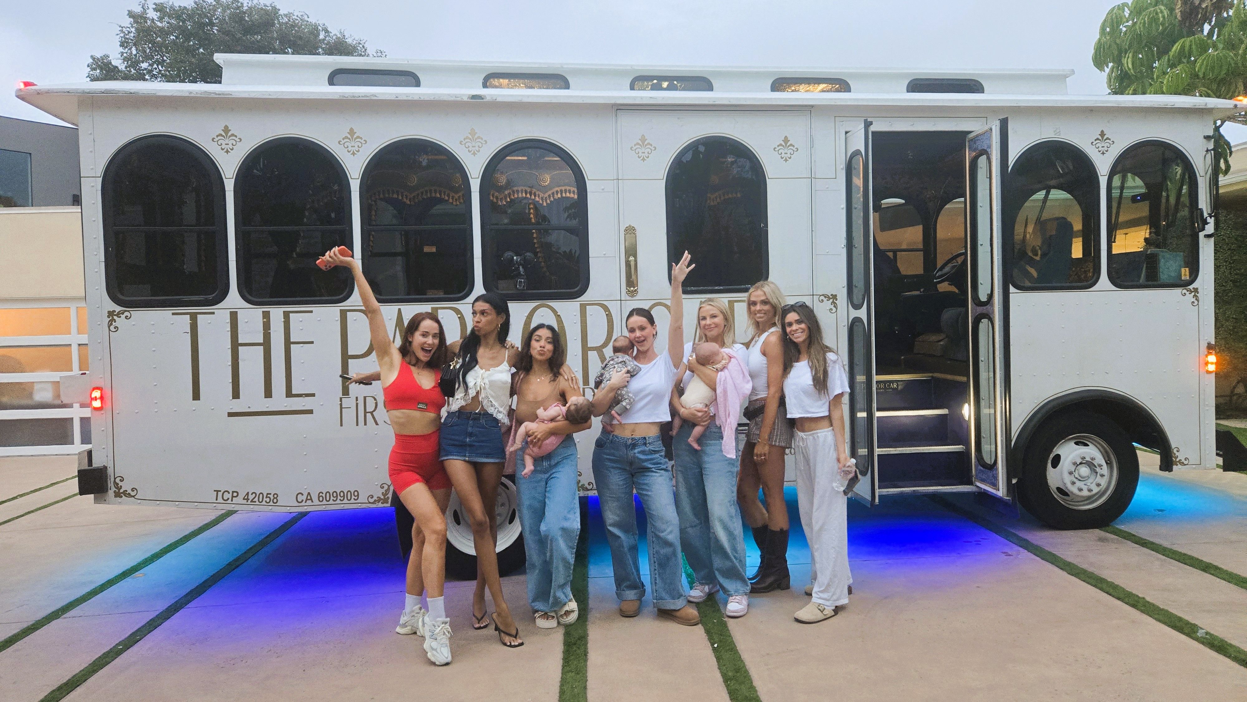 Group photo of seven women in casual outfits posing in front of a decorative white trolley with gold trim and blue underglow lights on a suburban driveway, several women holding babies and smiling