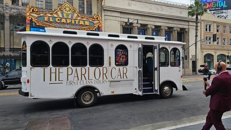 White vintage-style tour trolley labeled THE PARLOR CAR parked on a Los Angeles tourist street in front of an ornate theater marquee and classical stone building, while a suited cameraman films on the sidewalk.