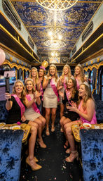 Bachelorette party on a luxury party bus — smiling women in tiaras and pink sashes singing karaoke, taking selfies amid confetti, drinks and snacks under an ornate blue-and-gold ceiling.