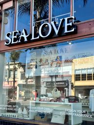 Sunlit coastal-chic storefront in Pasadena with a large illuminated sign, reflective glass showing palm trees and neighboring buildings, and a window display of pillows, candles and a woven bag.