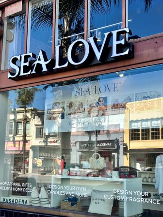 Sunlit coastal-chic storefront in Pasadena with a large illuminated sign, reflective glass showing palm trees and neighboring buildings, and a window display of pillows, candles and a woven bag.