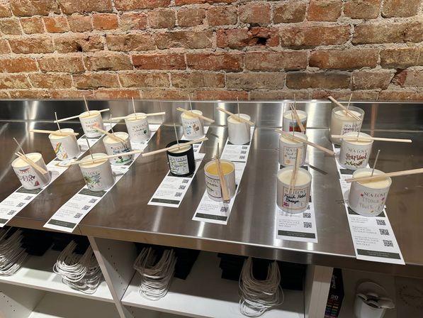 Candle scent sampling display on a stainless steel counter against an exposed brick wall, paper-cup testers with wooden scent sticks and labels