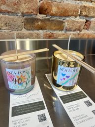 Cozy display of two hand-poured decorative candles in glass jars with wooden wick holders on a metal counter against an exposed brick wall, colorful labels and informational flyers with QR codes