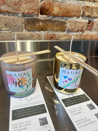 Cozy display of two hand-poured decorative candles in glass jars with wooden wick holders on a metal counter against an exposed brick wall, colorful labels and informational flyers with QR codes