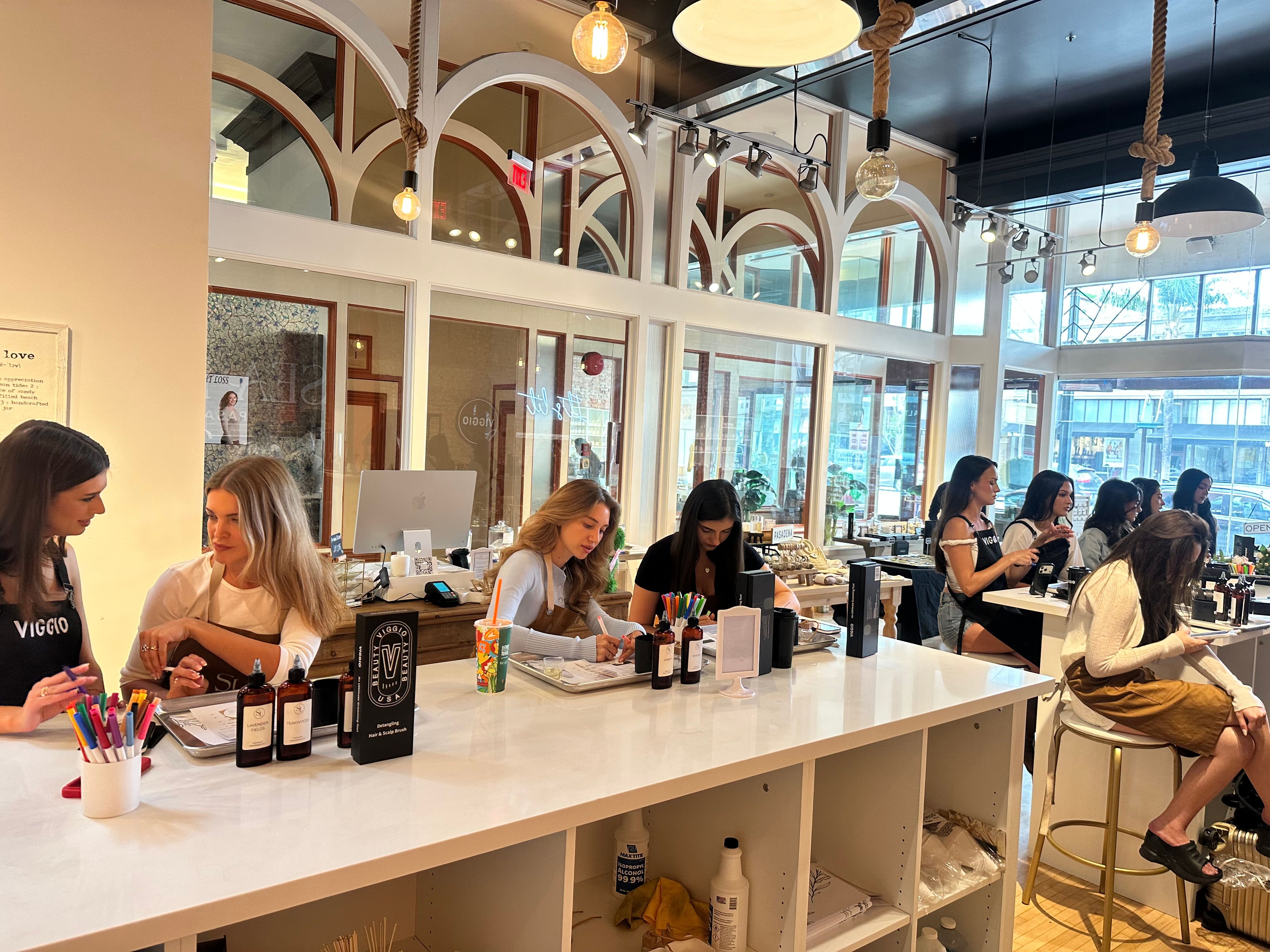Bright modern nail and beauty bar with arched interior windows and hanging pendant bulbs, long white counter where stylists attend clients on manicures and beauty services, large storefront windows with city view.