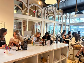 Bright modern nail and beauty bar with arched interior windows and hanging pendant bulbs, long white counter where stylists attend clients on manicures and beauty services, large storefront windows with city view.