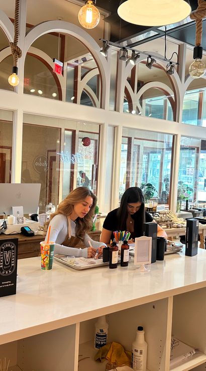 Bright modern nail and beauty bar with arched interior windows and hanging pendant bulbs, long white counter where stylists attend clients on manicures and beauty services, large storefront windows with city view.