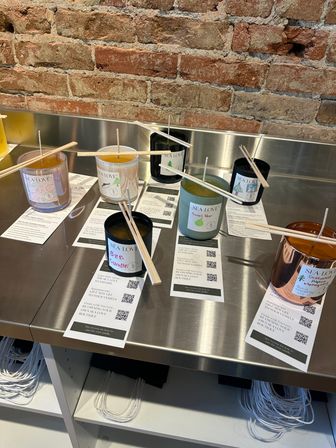 Artisan scented candles in assorted glass jars with wooden wick sticks arranged on a stainless-steel display table against an exposed brick wall in a boutique.
