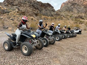 Six helmeted ATV riders lined up on quad bikes with a side-by-side in a rocky desert canyon on a gravel trail — ready for an off-road adventure.