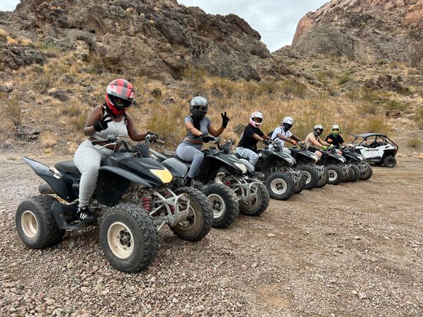 Six helmeted ATV riders lined up on quad bikes with a side-by-side in a rocky desert canyon on a gravel trail — ready for an off-road adventure.
