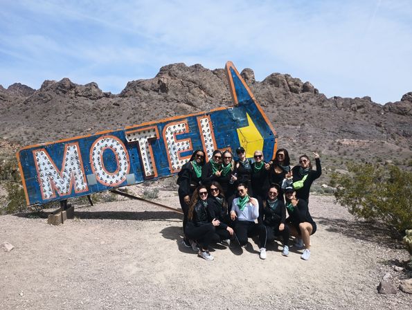 Group of people wearing green bandanas posing in front of a tilted vintage motel sign on a sunny roadside with rocky desert hills and blue sky