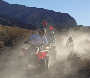 Three helmeted riders on red ATVs kicking up dust along a desert off-road trail with red safety flags and a rugged mountain backdrop under a clear blue sky.