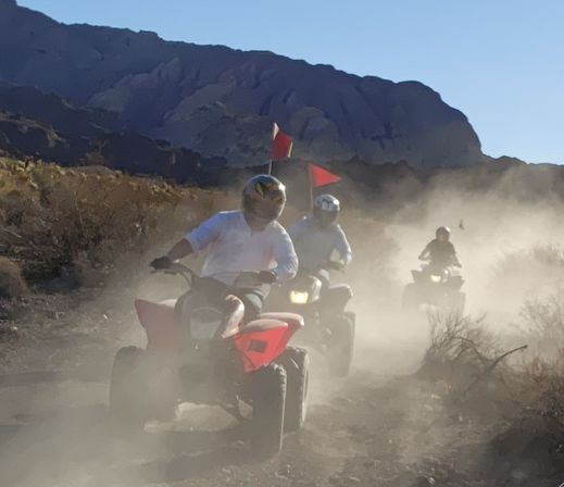Three helmeted riders on red ATVs kicking up dust along a desert off-road trail with red safety flags and a rugged mountain backdrop under a clear blue sky.