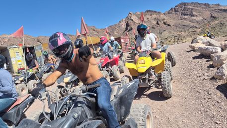 Group of helmeted riders on ATVs lined up on a dusty desert trail for an off-road ATV tour, rocky canyon backdrop, red safety flags and clear blue sky