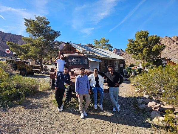 Five men posing on a rusty vintage truck in a sunlit desert ghost‑town setting with a weathered wooden cabin, scrubby desert plants and rocky mountains under a bright blue sky.