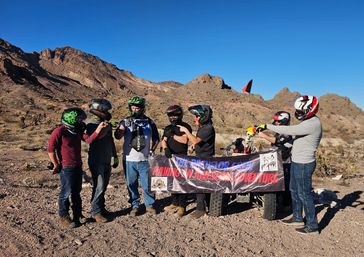 Helmeted off-road riders holding a banner beside an ATV with a skeleton passenger in a rocky desert with rugged mountains and clear blue sky — desert ATV adventure
