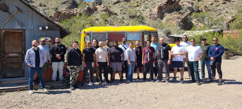 Team outing group photo of about 20 men posing in front of a weathered yellow school bus and corrugated metal cabin in a sunny desert canyon with rocky hills and gravel foreground
