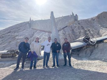 Five people posing in front of a weathered airplane wreck on a sunlit arid rocky hillside, with broken fuselage and wings half-buried in the white textured terrain.