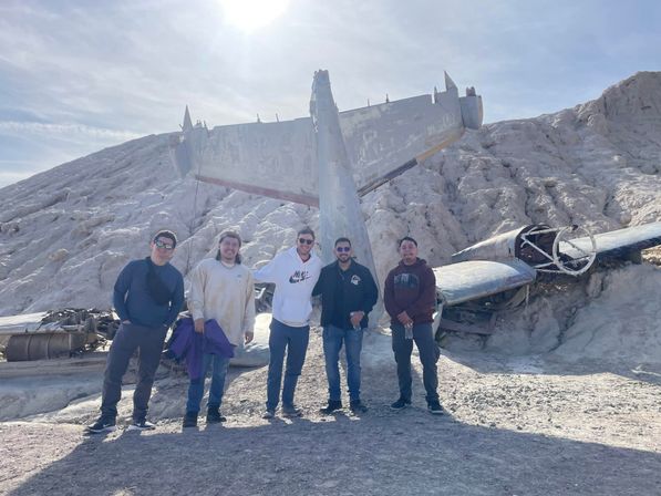Five people posing in front of a weathered airplane wreck on a sunlit arid rocky hillside, with broken fuselage and wings half-buried in the white textured terrain.