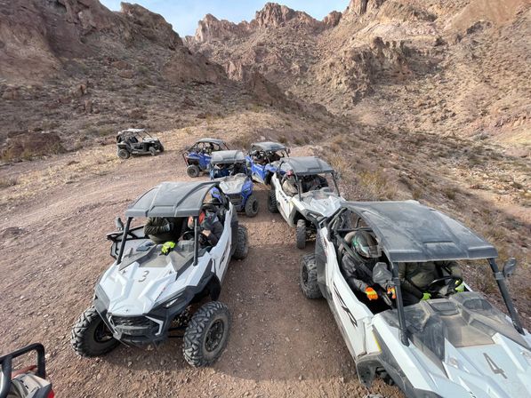 Line of side-by-side UTVs with helmeted riders parked on a dusty desert canyon trail beneath rugged rocky cliffs