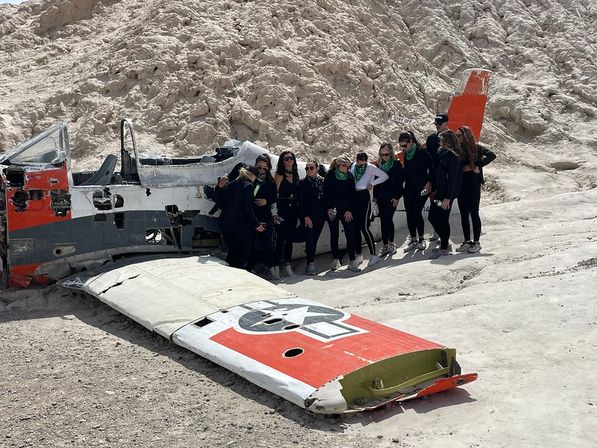 Adventurers posing beside a weathered orange-and-white military plane fuselage and detached wing with a roundel, stranded on a pale rocky desert slope.