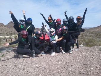 Group of ATV riders in colorful helmets and bandanas raising their arms on a red quad in a rocky desert under a bright blue sky