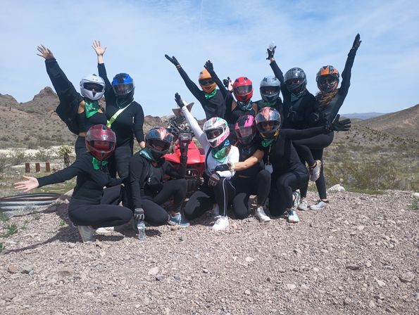 Group of ATV riders in colorful helmets and bandanas raising their arms on a red quad in a rocky desert under a bright blue sky