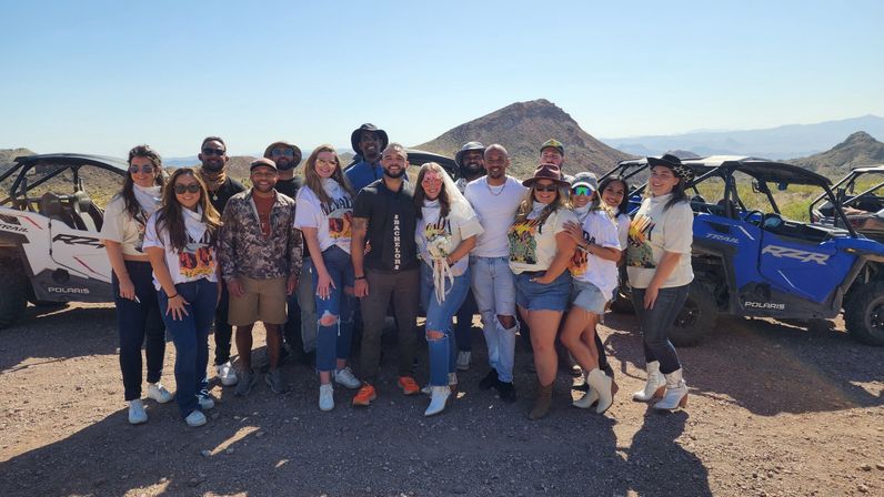 Group of friends in casual western outfits posing by off-road UTVs on a sunny desert trail with rocky hills in the background; one person wears a veil and holds a bouquet.