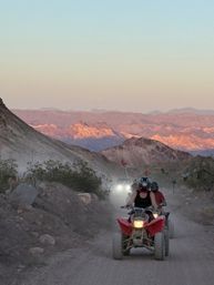 ATV riders on a dusty desert trail at sunset, helmets and red safety flags visible, headlights cutting through dust with rugged pink-hued mountains in the distance.