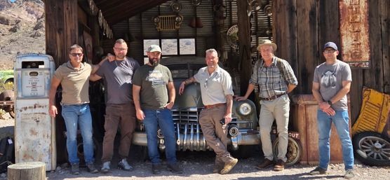 Six men posing in front of a vintage mid-century car inside a rustic desert garage with a weathered gas pump, antique parts, and rocky hills in the background.