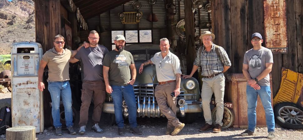 Six men posing in front of a vintage mid-century car inside a rustic desert garage with a weathered gas pump, antique parts, and rocky hills in the background.
