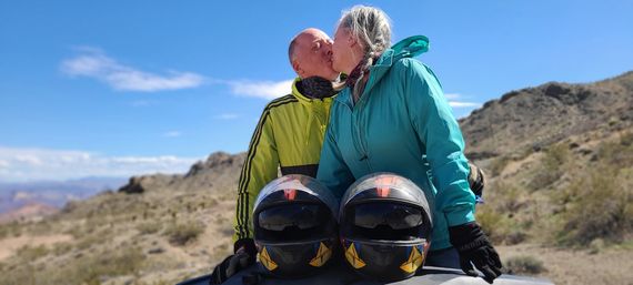 Couple kissing beside two motorcycle helmets on a vehicle hood in a sunny desert landscape with blue sky and rocky hills.