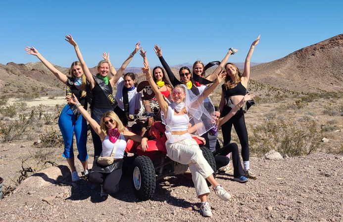 Group of friends in sashes, bandanas and a veil posing with arms raised on an ATV during a sunny bachelorette desert outing amid rocky hills and clear blue sky.