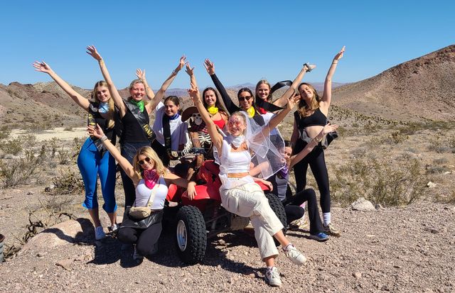 Group of friends in sashes, bandanas and a veil posing with arms raised on an ATV during a sunny bachelorette desert outing amid rocky hills and clear blue sky.