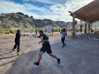 Three people in athletic clothes playfully chase each other with water guns near a wooden pavilion in a sunlit desert mountain landscape
